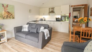 A modern living room with a grey sofa, wooden dining table with chairs, a kitchen with white cabinets, and a painting of a rabbit on the wall. - Sunrise Stable (Spalding)
