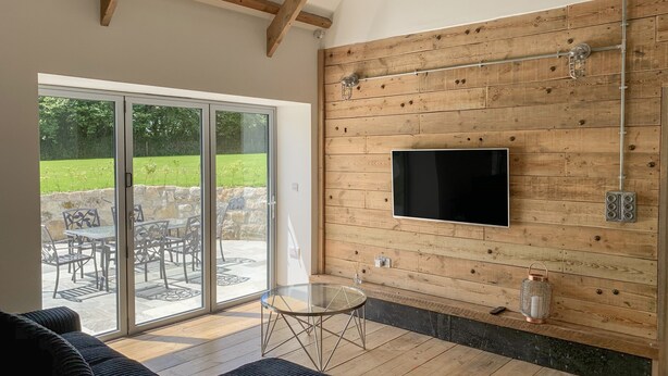 Una sala de estar moderna con un televisor grande montado en una pared de madera, un amplio ventanal corredero que da a un patio exterior y una zona de estar acogedora con sofá y mesa de vidrio.