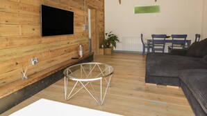 Una habitación con paredes de madera, un televisor de pantalla plana montado en la pared, una mesa de vidrio con patas de metal, un sofá oscuro y una zona de comedor con sillas azules y una mesa.