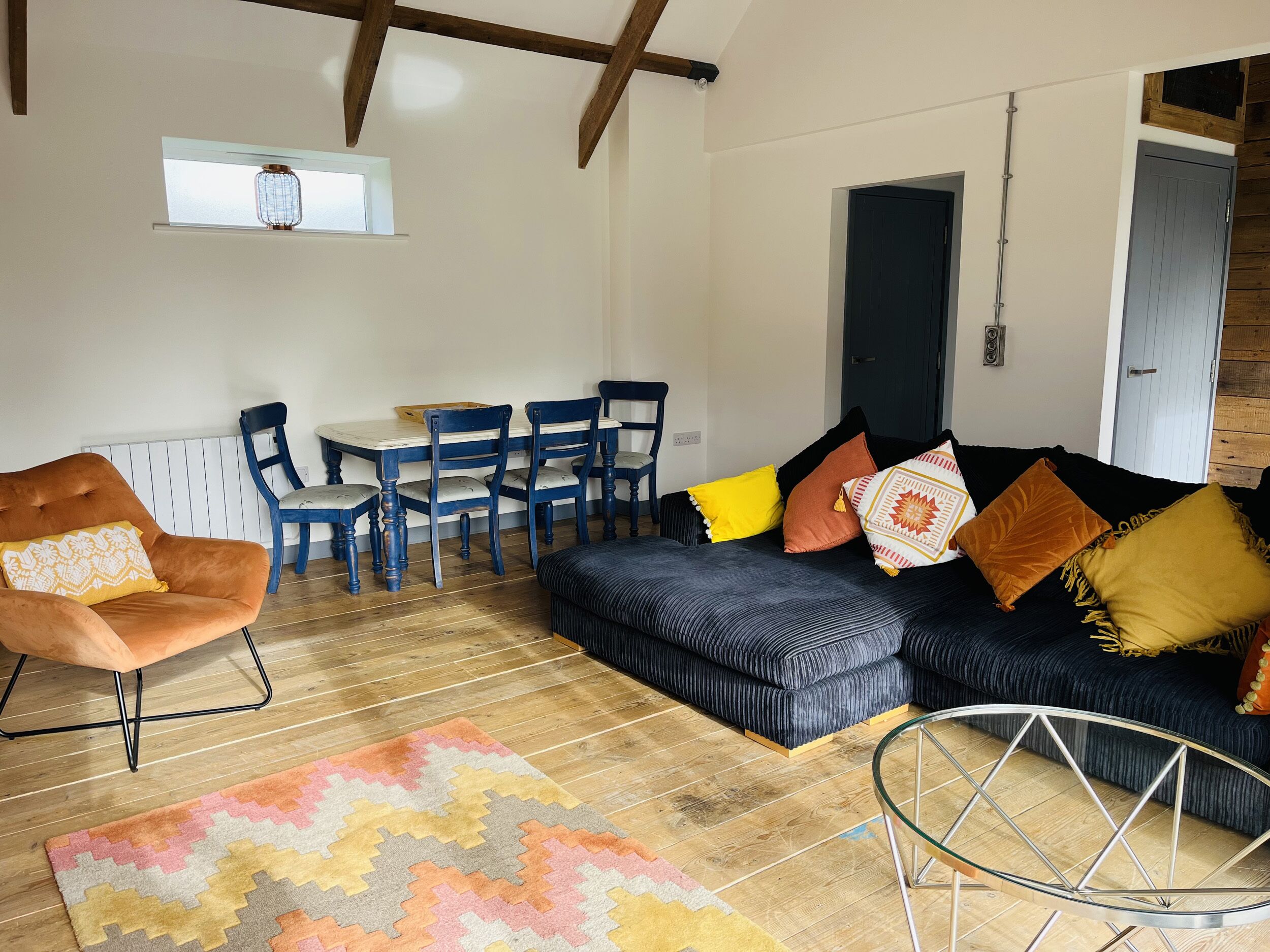 A cozy living room with a dark blue sofa, a wooden dining table with blue chairs, and a patterned rug.