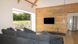 Una sala de estar moderna con un sofá gris, un televisor de pantalla plana montado en una pared de madera, un muro de piedra visible a través de un vidrio y un refrigerador de acero inoxidable al fondo.