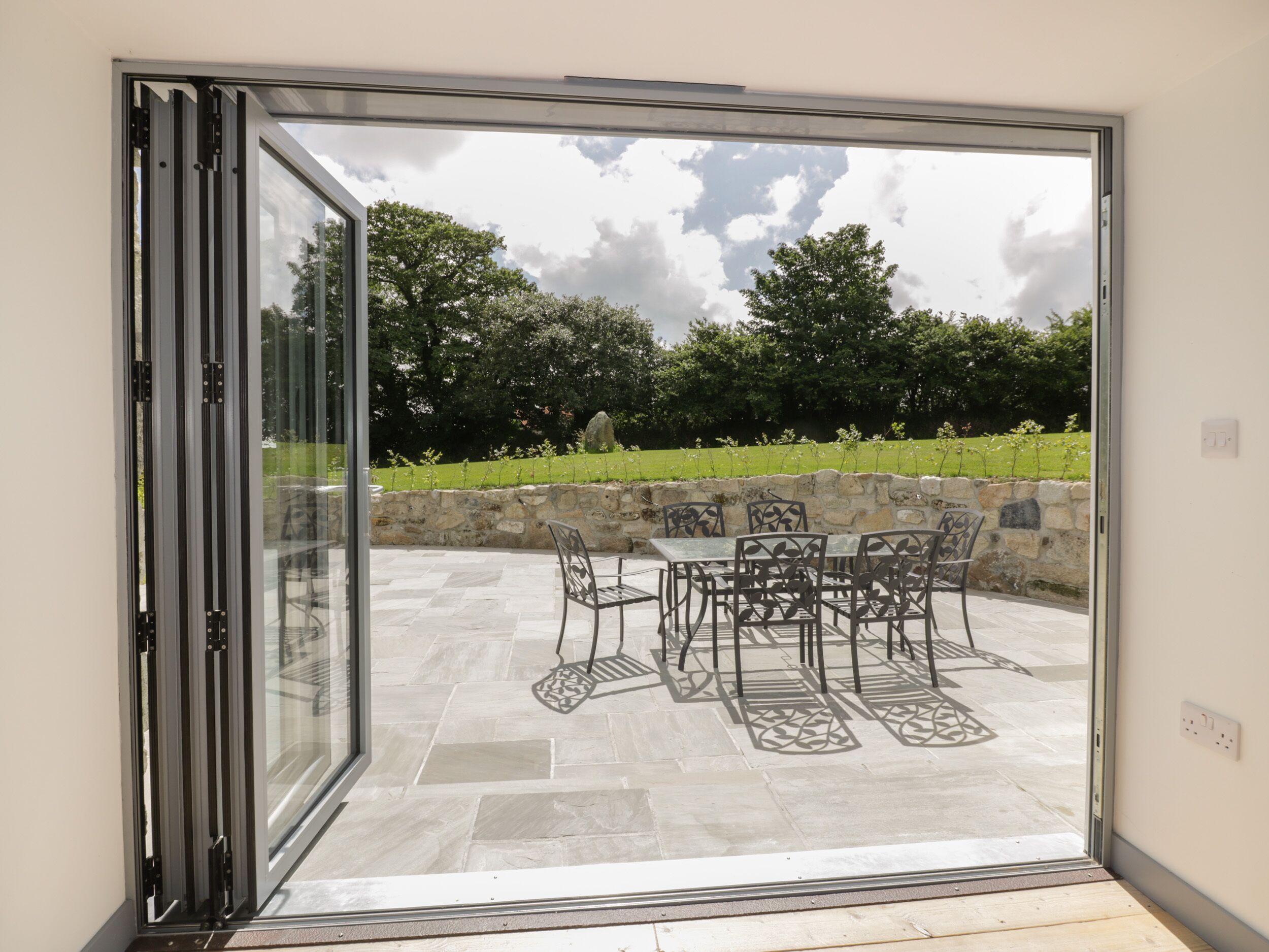 A patio with a table and chairs, a stone wall, and a view of trees and grass.