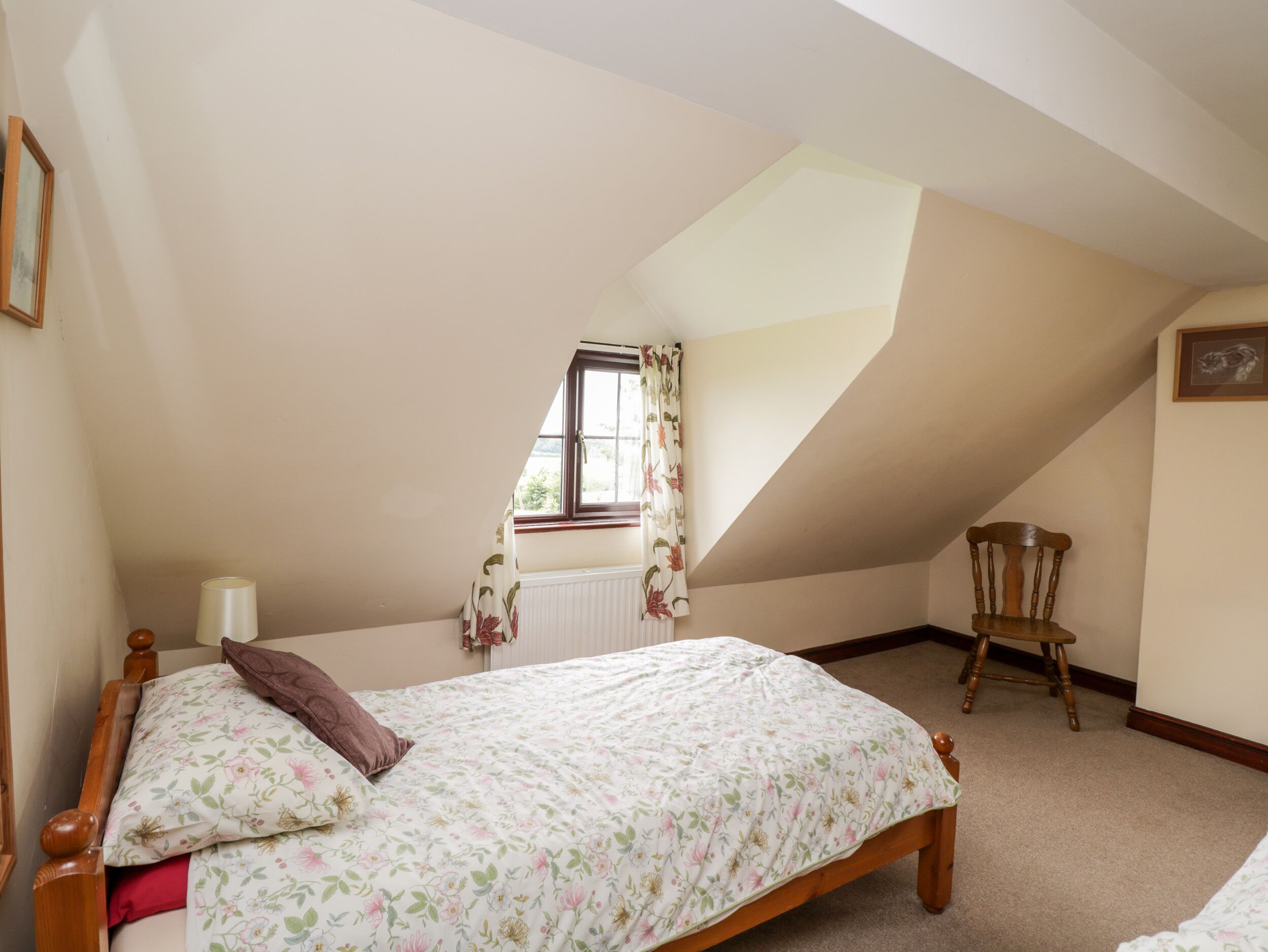 A bedroom with a sloped ceiling, a wooden bed, a chair, a window with floral curtains, and a radiator.