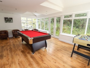 A pool table with a red felt top, two brown armchairs, and a foosball table in a room with large windows and wooden flooring.