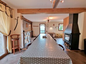 A long dining table with chairs, a kitchen area with a wood stove, stone wall decorations, and a view through a window.