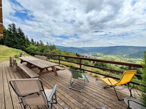 Een houten terras met een picknicktafel en stoelen, met uitzicht op een vallei met bergen en een stad.