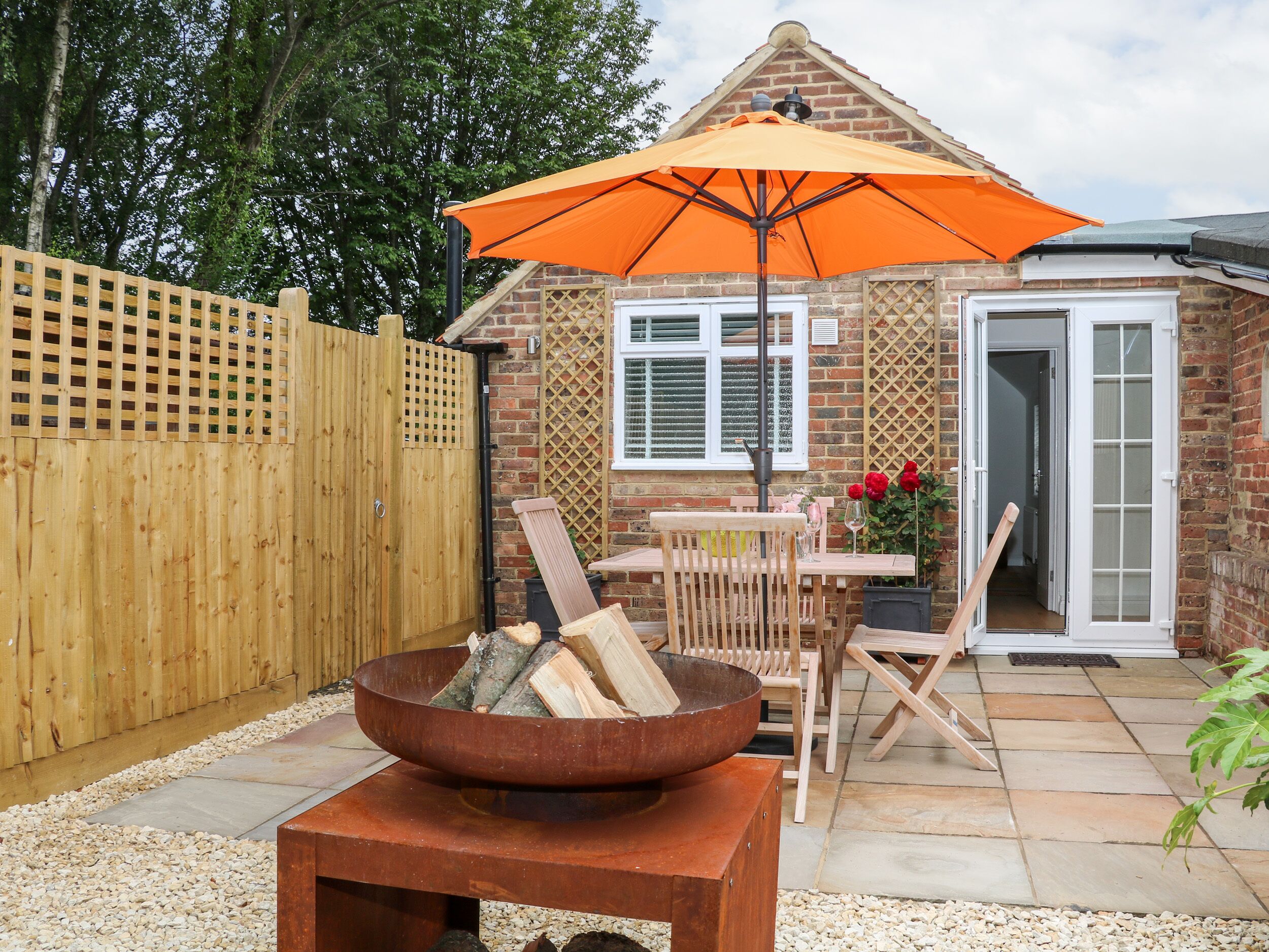 A patio area with a wooden table, chairs, a fire pit, and an orange umbrella.