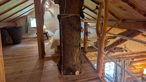 Een rustiek houten interieur met een schuine balkenkap, een grote boomstam en een touwladdertje.
