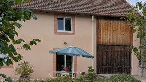 A two-story house with a tiled roof, a wooden door, and a small patio area with a table and chair. - Spacious Gîte: Fireplace, Terrace, Near Epinal & Gérardmer, Sleeps 5 (Docelles)