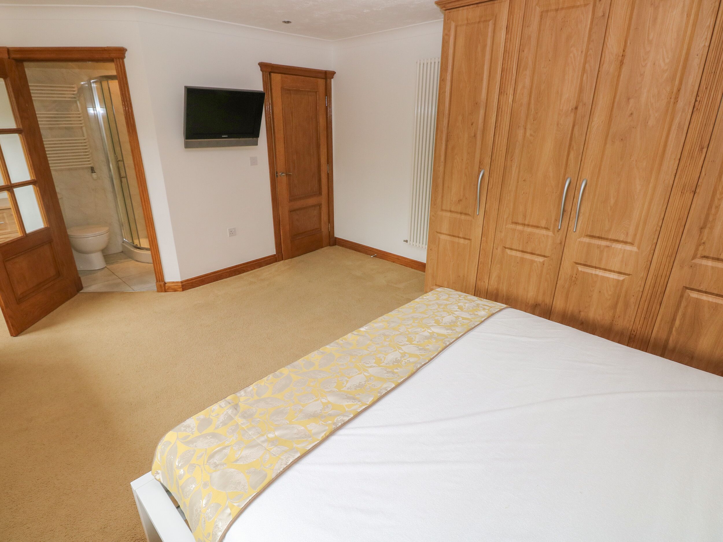 Un dormitorio con cama, armario de madera, televisor en la pared y baño con inodoro y ducha.