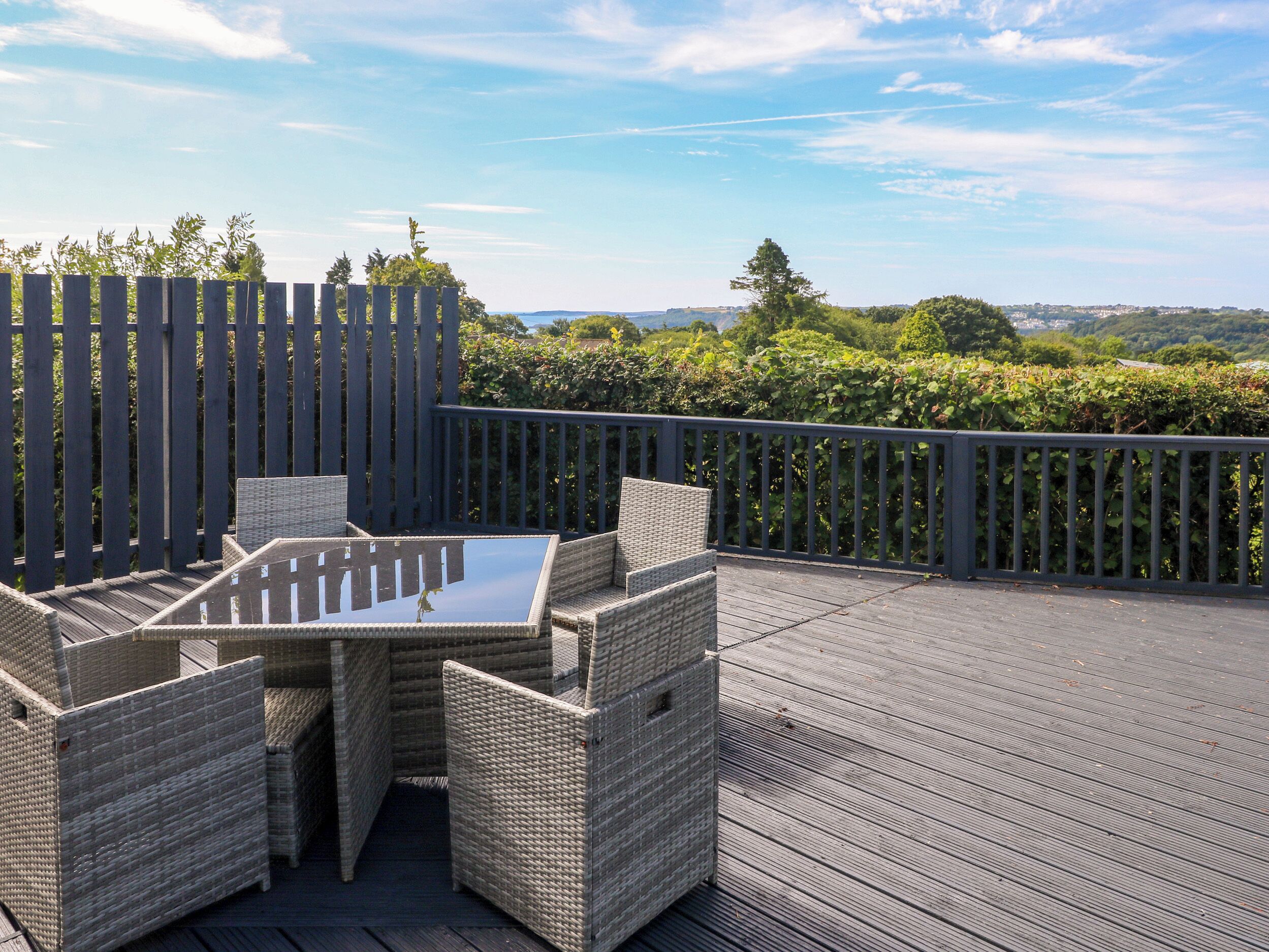 An outdoor deck with wicker furniture and a glass-top table, surrounded by a wooden fence and lush greenery.