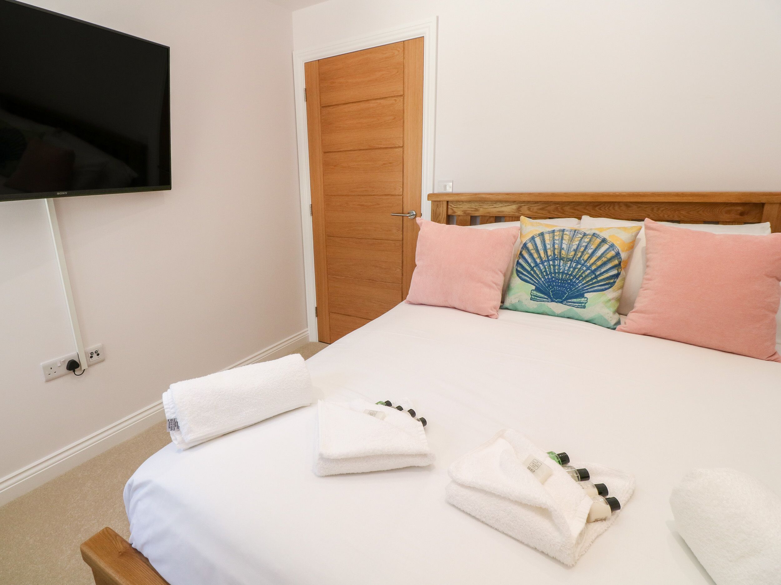 A neatly made bed with white linens, a wooden headboard, and a flat-screen TV mounted on the wall.