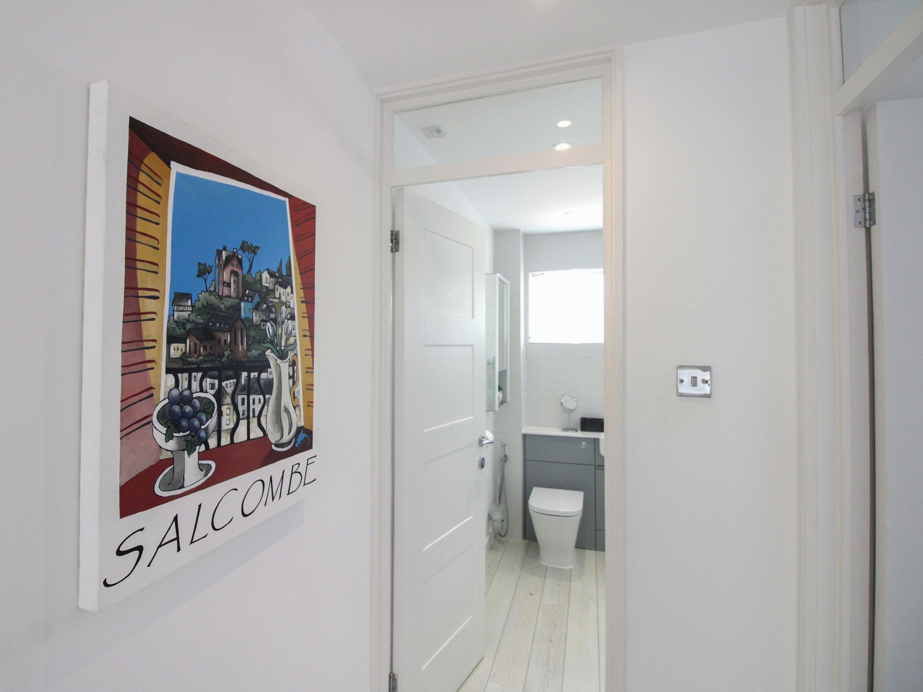 A hallway leading to a bathroom with a white door, a framed artwork on the wall, and a visible toilet.