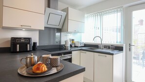 A modern kitchen with a black countertop, white cabinets, a built-in oven, and a window with blinds. - Little Starboard (Kingsbridge)