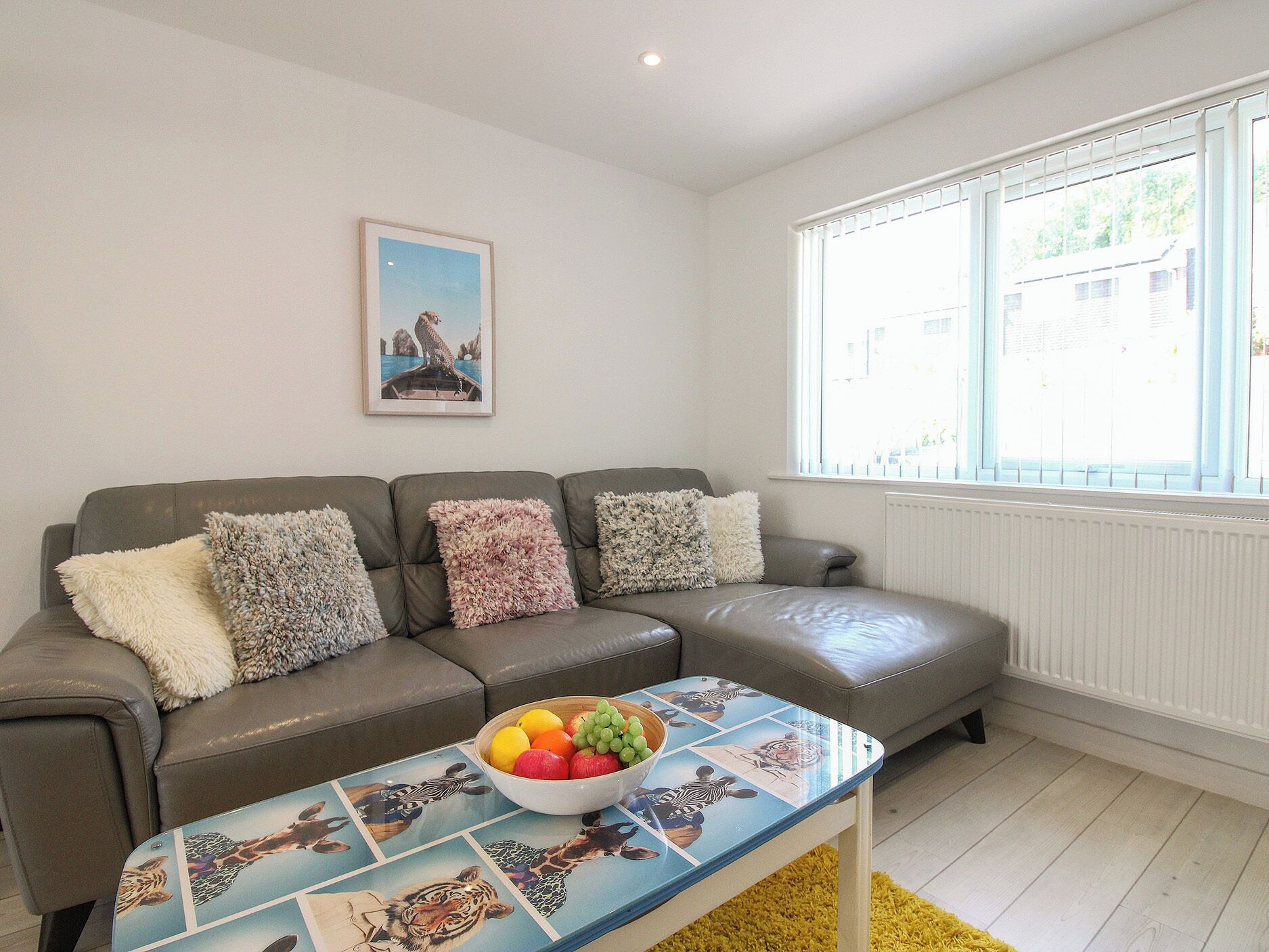 A living room with a grey sofa, a coffee table with a fruit bowl, and a window with blinds.