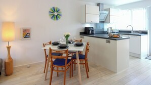 A modern kitchen with white cabinets, a central island, and a dining area with wooden chairs and a table. - Little Starboard (Kingsbridge)
