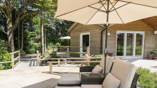 A log cabin with a patio area featuring wicker furniture and a large umbrella. - Brambleside Lodge (Truro)