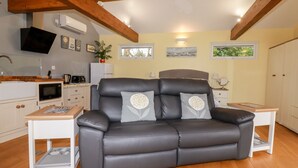 A modern living room with a grey leather sofa, wooden beams, a kitchen area, and a dining table. - Brambleside Lodge (Truro)