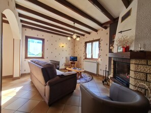 Ein Wohnzimmer mit Sofa, Sessel, Fernseher, Kamin und Blick ins Freie.