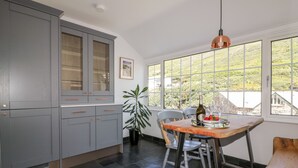 A modern kitchen with grey cabinets, a dining table with chairs, a potted plant, and a view of greenery outside. - Harbour Cottage (Boscastle)