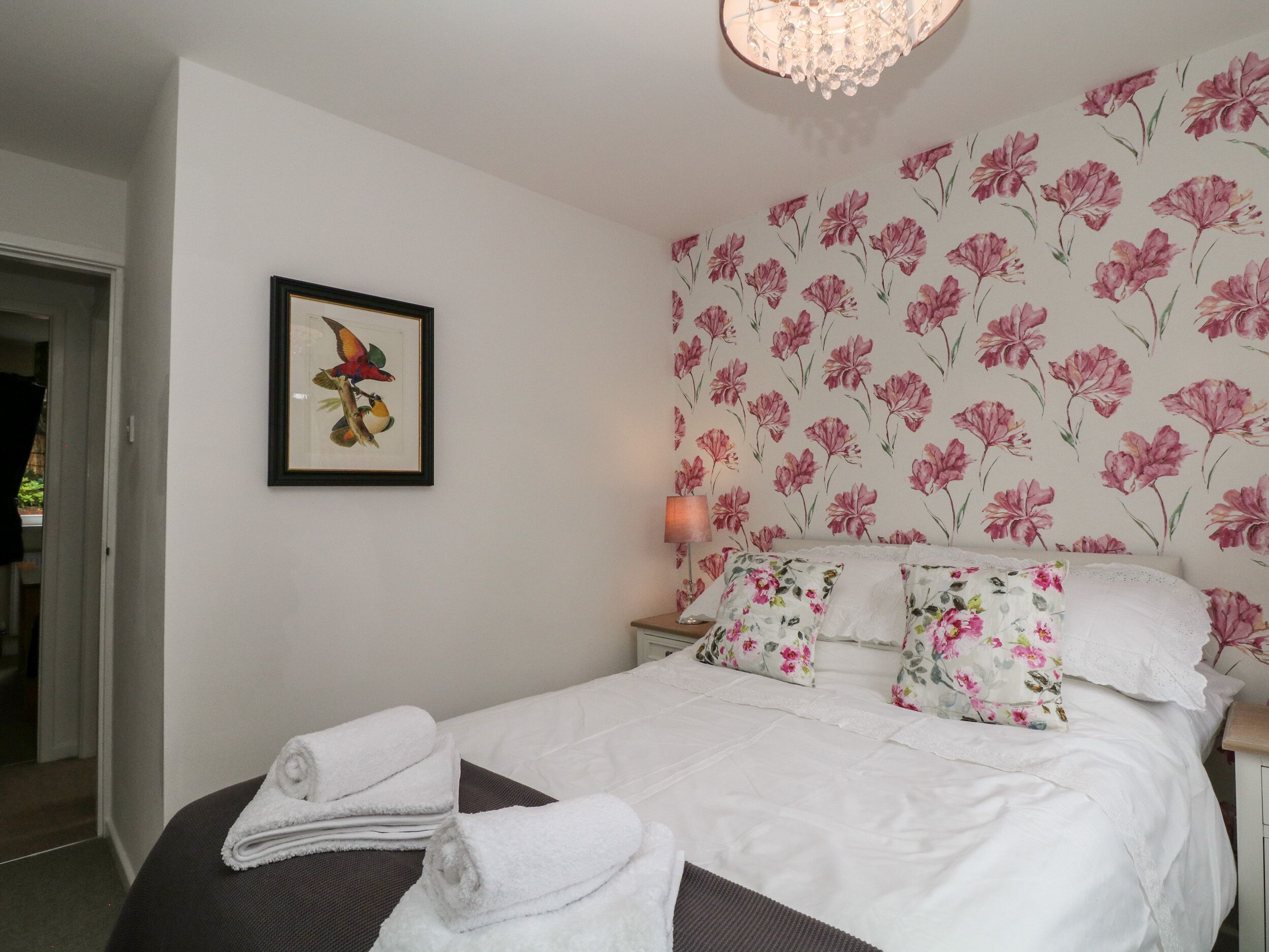 A neatly made bed with floral pillows, a framed artwork of birds, and a floral wallpaper.