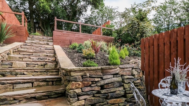 A stone staircase leading to a garden area with a wooden fence, a metal table with chairs, and a variety of plants. - Hunters Cottage (High Peak)