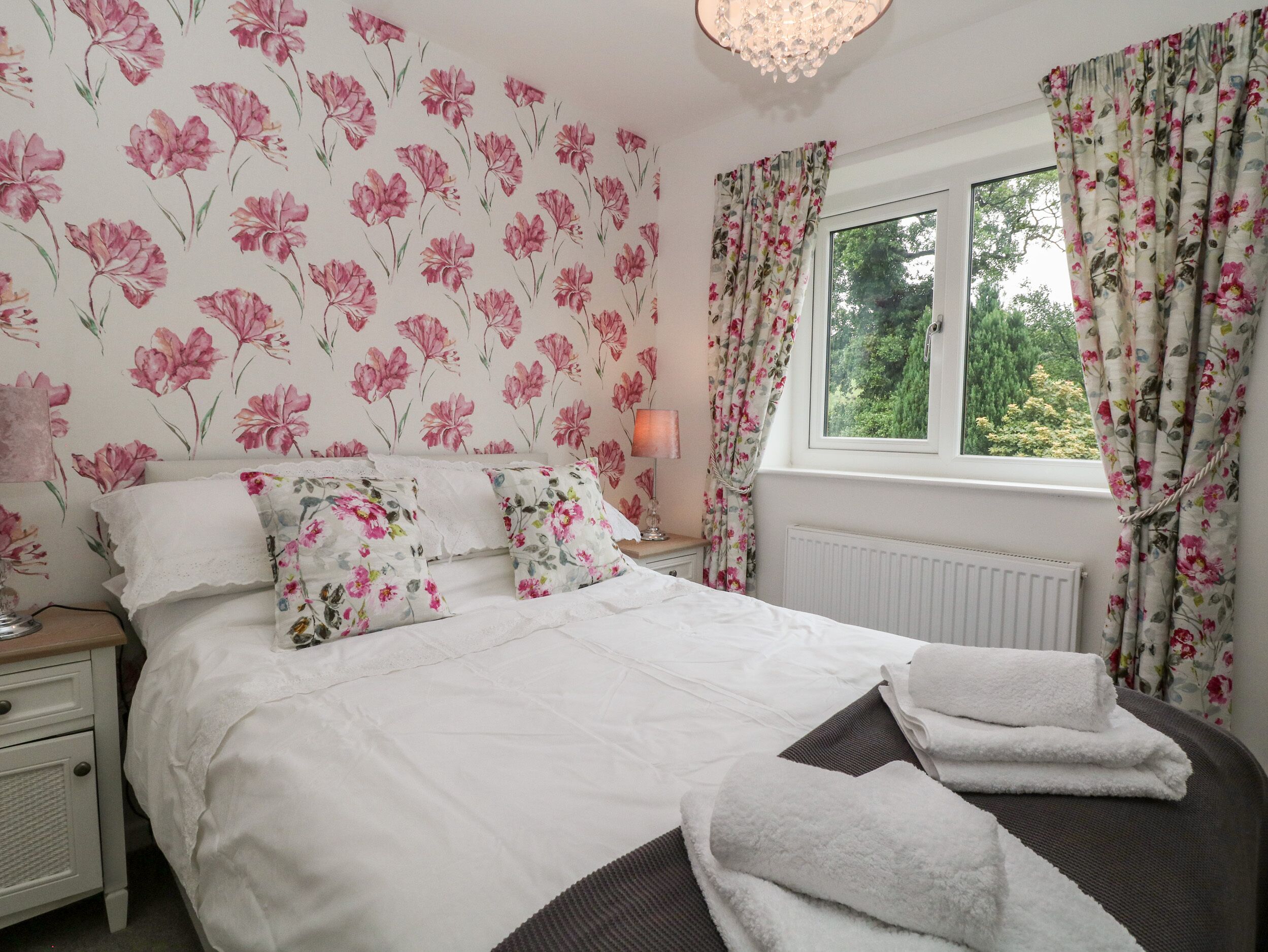 A bedroom with floral wallpaper, a bed with white linens, a bedside table with a lamp, and a window with curtains.