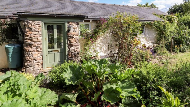Un rustico cottage in pietra con una porta verde, un piccolo annesso e un giardino con piante a foglia grande.