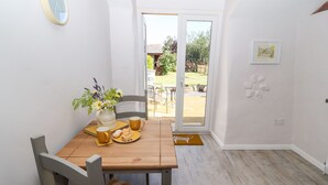 Zona pranzo con tavolo in legno, sedie, un vaso con fiori e un piatto con cibo.