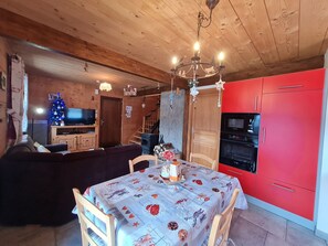 Uma sala de estar aconchegante com teto de madeira, uma mesa de jantar com toalha festiva, uma cozinha vermelha e uma televisão ao fundo.