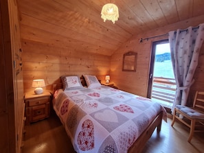 Quarto de cabana de madeira com cama, abajures, uma cadeira de madeira e vista para o exterior.