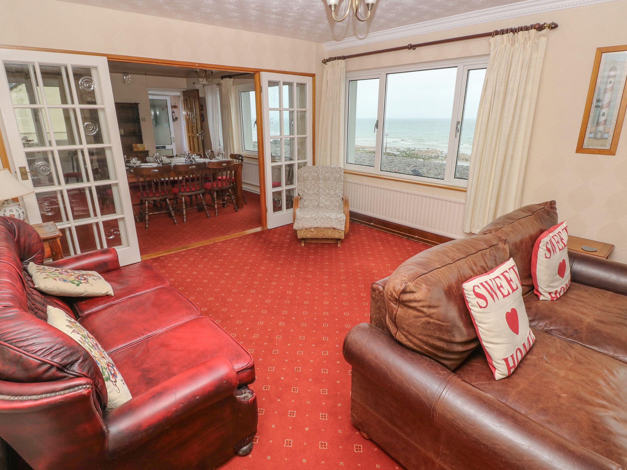 A living room with red carpet, a brown leather sofa, and a red leather armchair. There is a dining area visible through an open door.