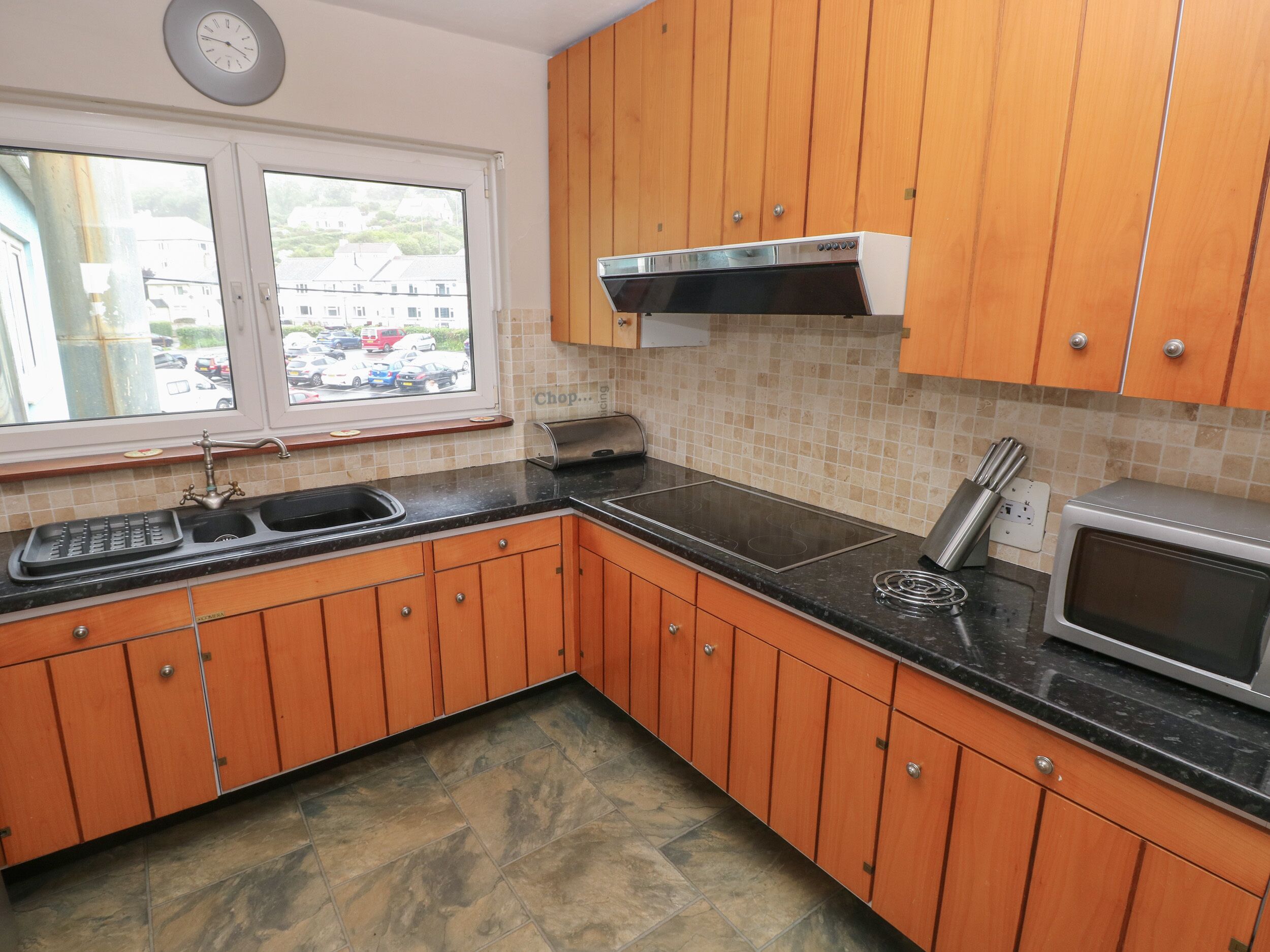 A kitchen with wooden cabinets, a black countertop, a stainless steel range hood, a microwave, and a window with a view of buildings outside.