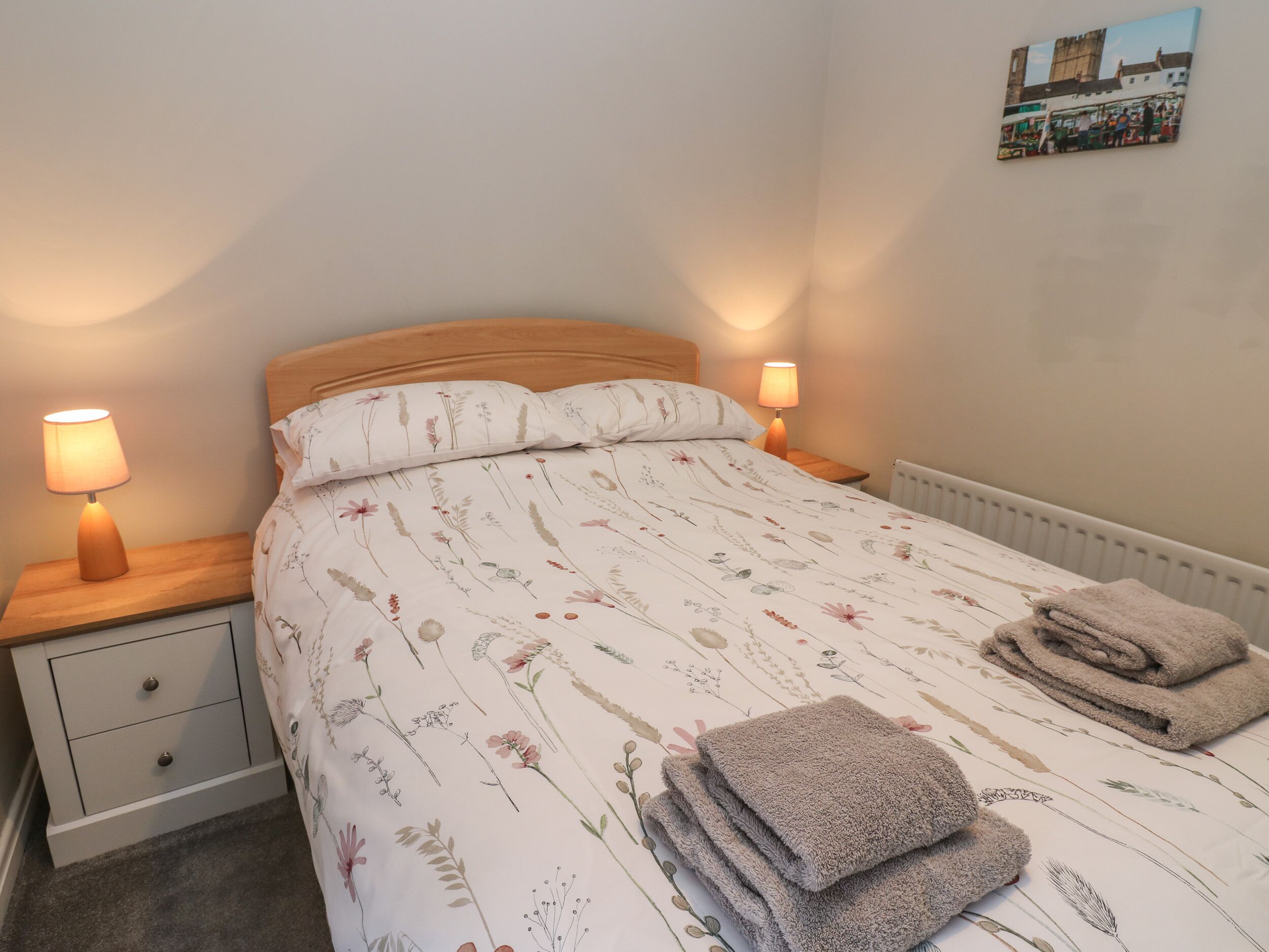 A neatly made bed with floral bedding, two bedside lamps, a small bedside table with a lamp, and a folded towel on the bed.