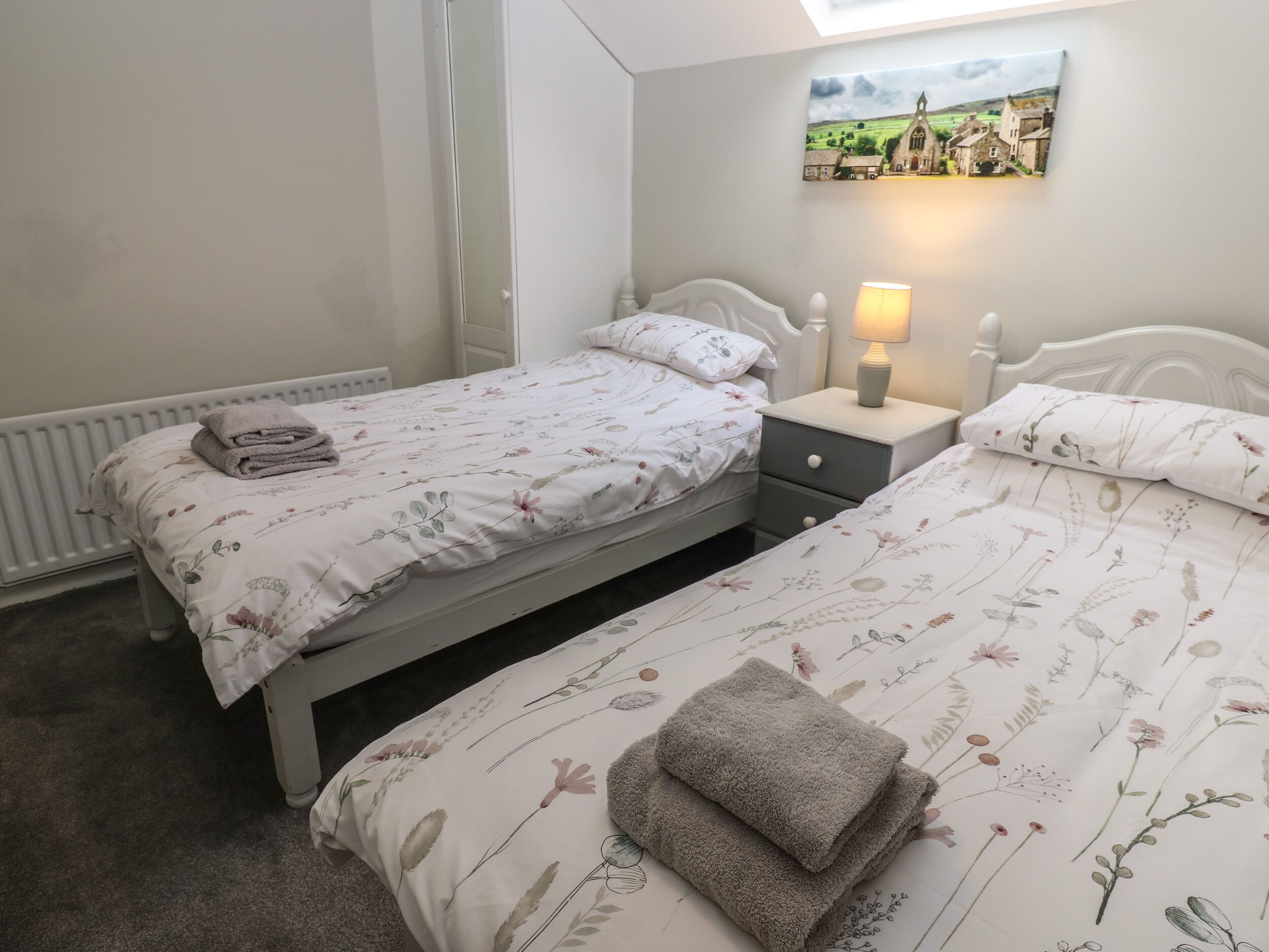 Quarto com duas camas de solteiro, uma mesa de cabeceira com abajur, um radiador e um quadro na parede.