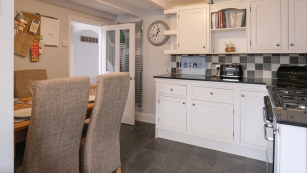A compact kitchen with white cabinets, a stove, and a dining area with two chairs and a table. - Lantern Cottage (St Austell)