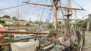 A historic sailing ship docked at a harbor with other boats, wooden deck, and a backdrop of buildings and greenery. - Lantern Cottage (St Austell)