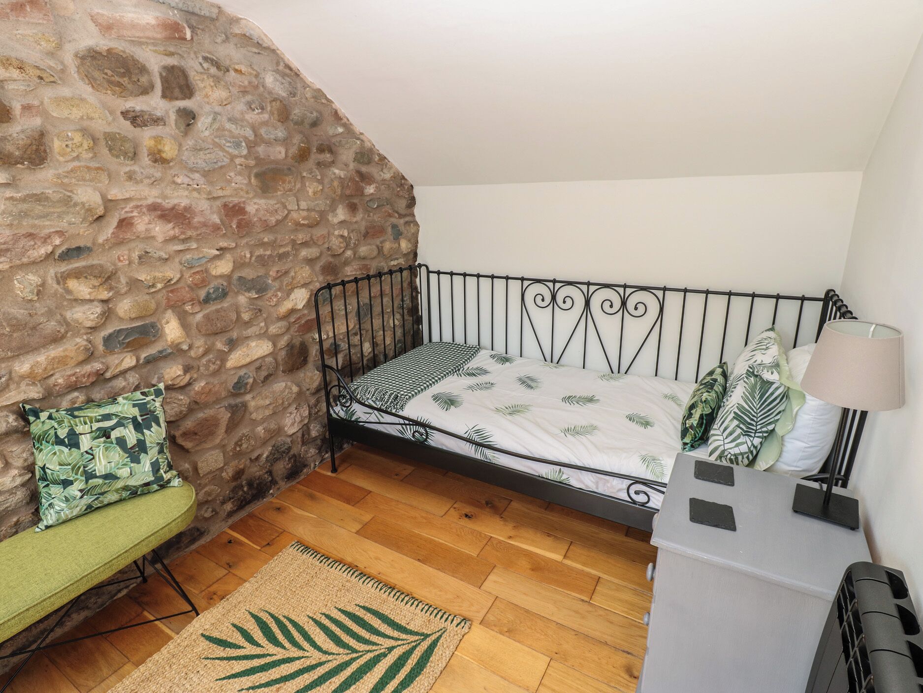 Una habitación pequeña y acogedora con una pared de piedra, estructura de cama de metal, una silla verde y piso de madera.