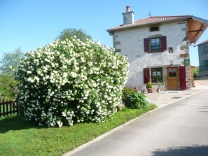 A stone building with a red door and shutters, surrounded by lush greenery and white flowers. - Cosy Cottage with Fireplace, Terrace & River View - Near Vittel Spa Towns (Bonvillet)