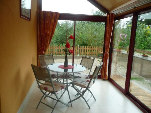 A conservatory with a round table and chairs, a wooden fence, and a garden view. - Cosy Cottage with Fireplace, Terrace & River View - Near Vittel Spa Towns (Bonvillet)