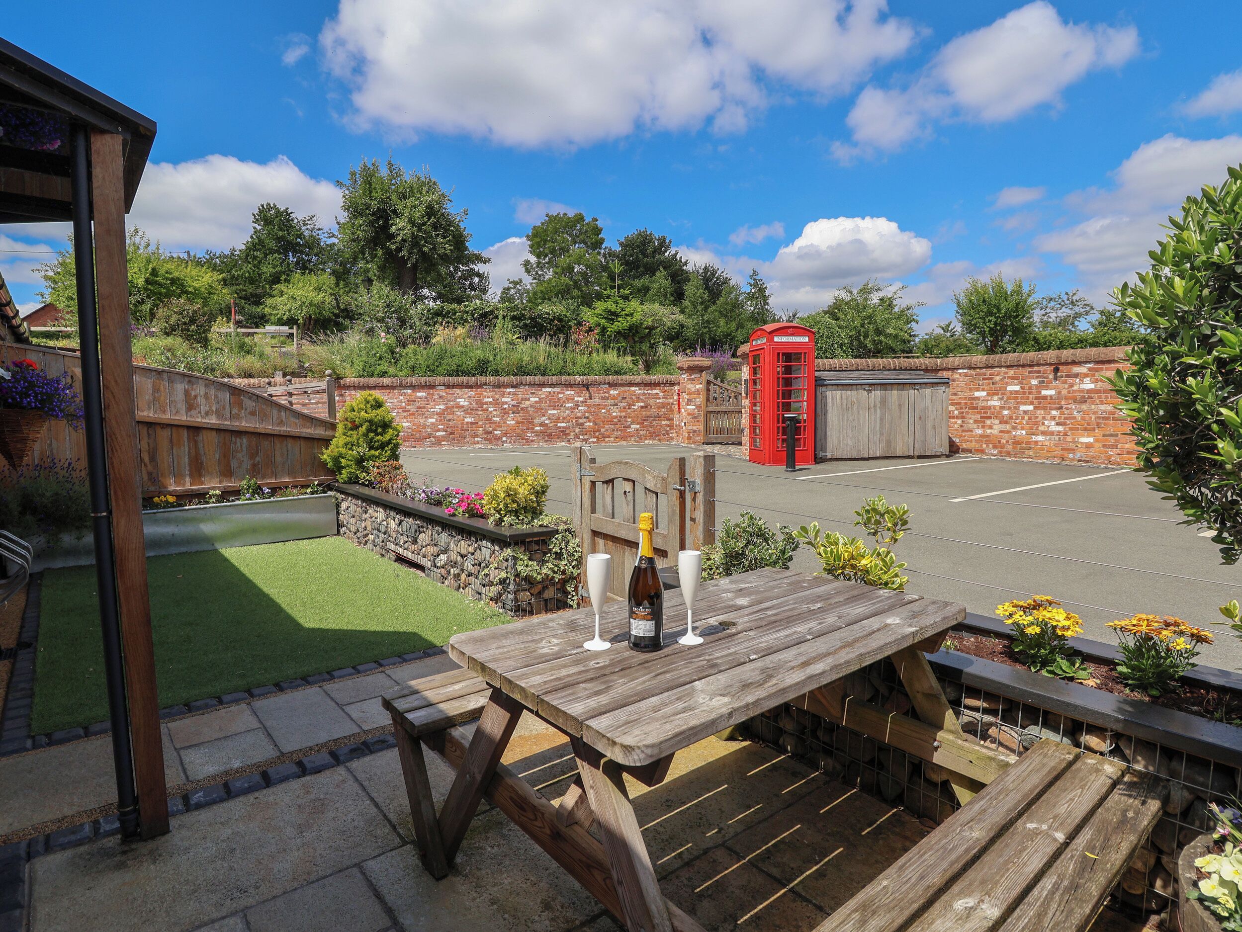 A wooden picnic table with a bottle and glass, surrounded by a garden with a brick wall and a red telephone booth in the background.