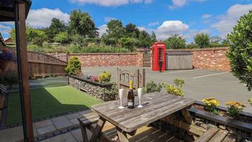 A wooden picnic table with a bottle and glass, surrounded by a garden with a brick wall and a red telephone booth in the background.
