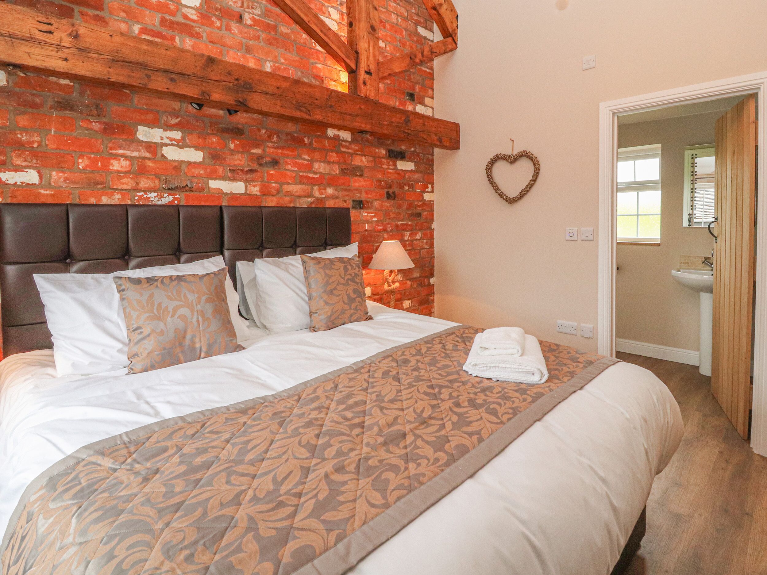 Una habitación de hotel con pared de ladrillo, cama con ropa de cama estampada, decoración con forma de corazón y baño visible a través de una puerta abierta.