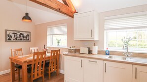 A modern kitchen with wooden dining table and chairs, white cabinets, and a window with blinds. - The Shippon (Chester)