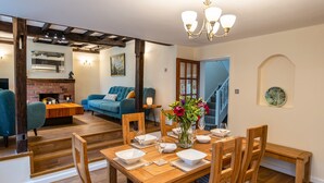 A dining area with a wooden table set for four, flanked by wooden chairs. A vase with flowers is on the table, and a staircase is visible in the background leading to another room. - Eleri Cottage (Malvern)