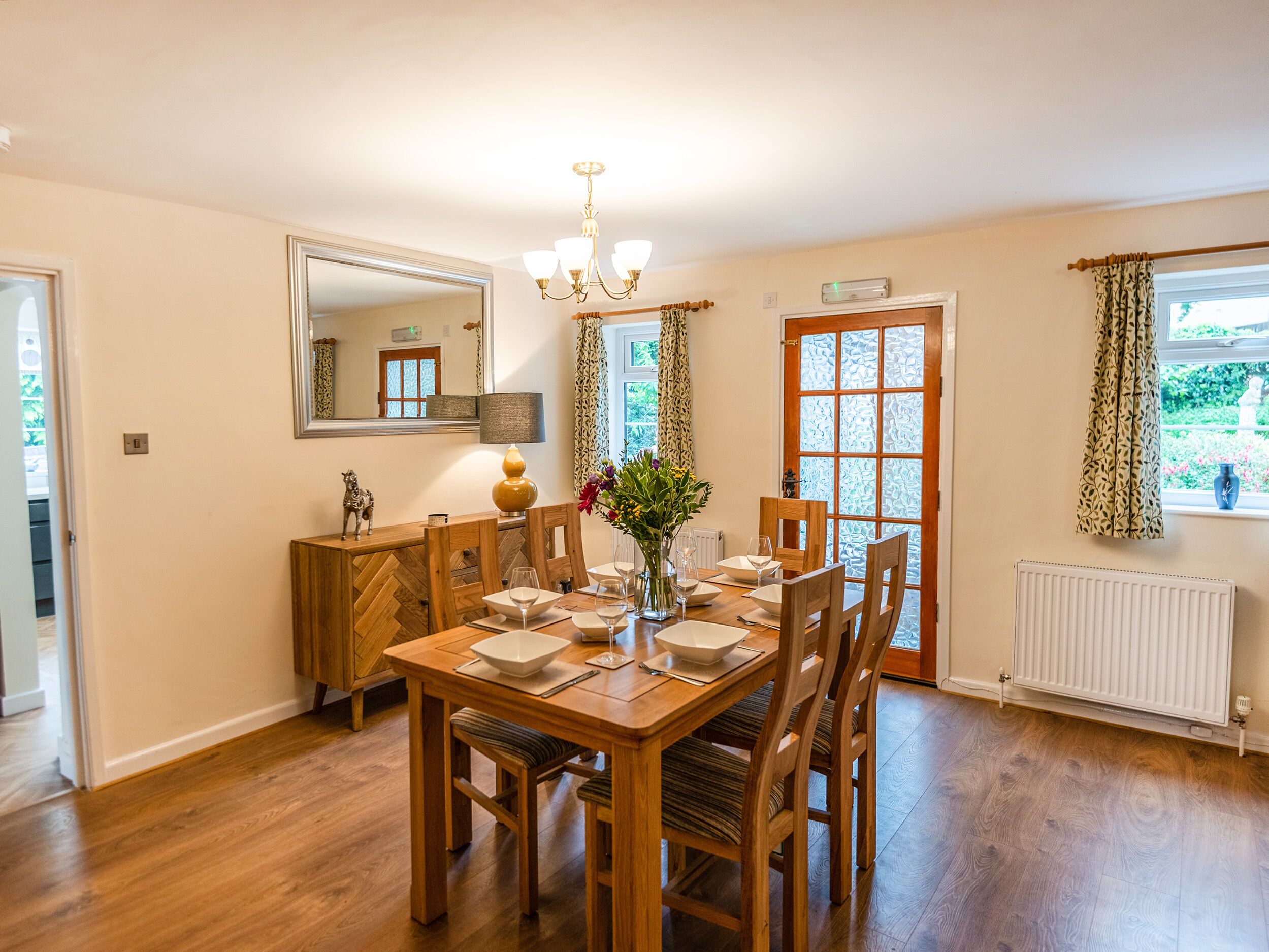 A dining room with a wooden table set for four, a chandelier, and a large window with patterned curtains.