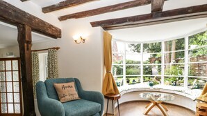 A cozy room with a teal armchair, a wooden table, and a bay window with a garden view. - Eleri Cottage (Malvern)
