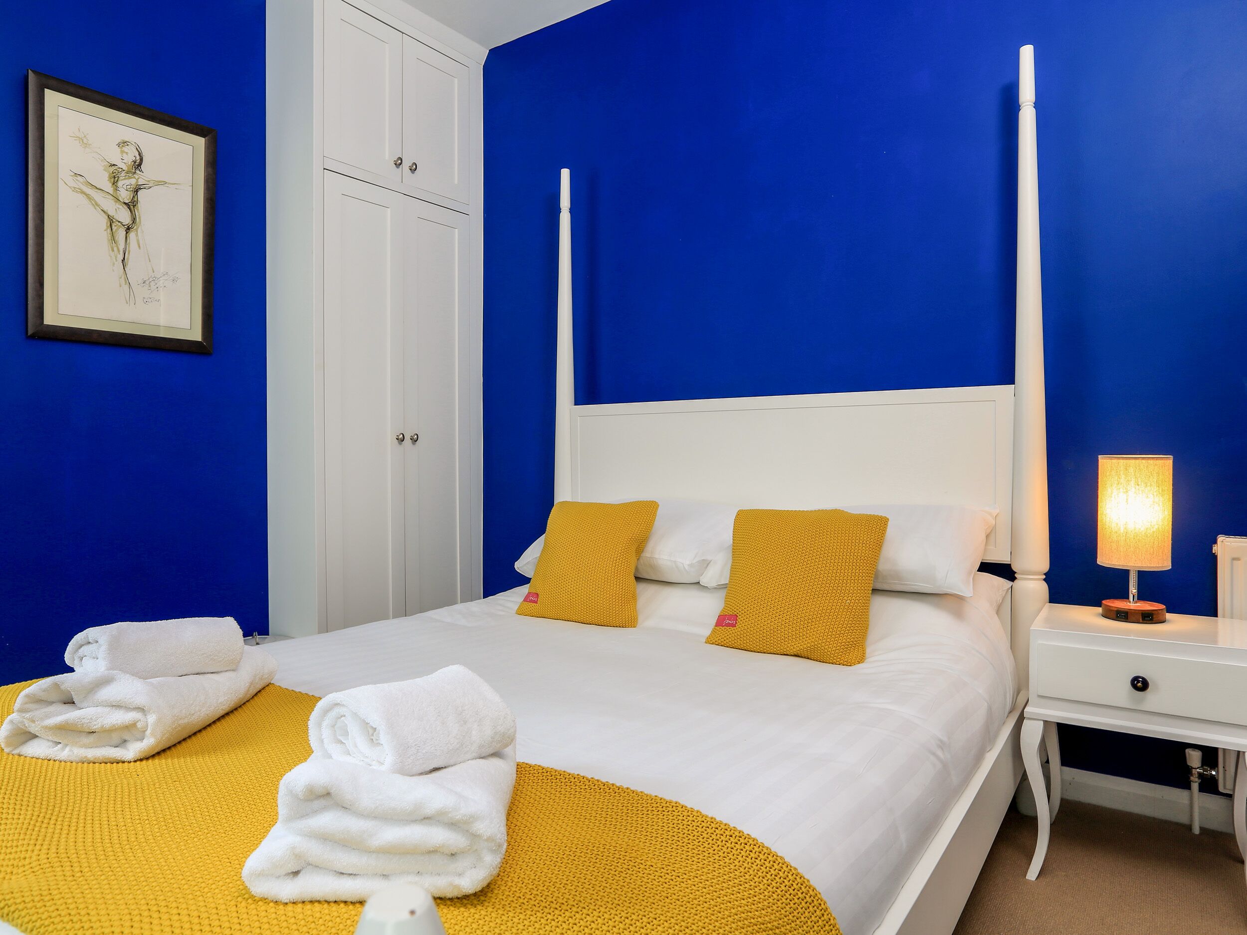 A bedroom with a four-poster bed, white bedding, and yellow accents. There is a framed artwork on the wall, a white wardrobe, and a bedside table with a lamp.