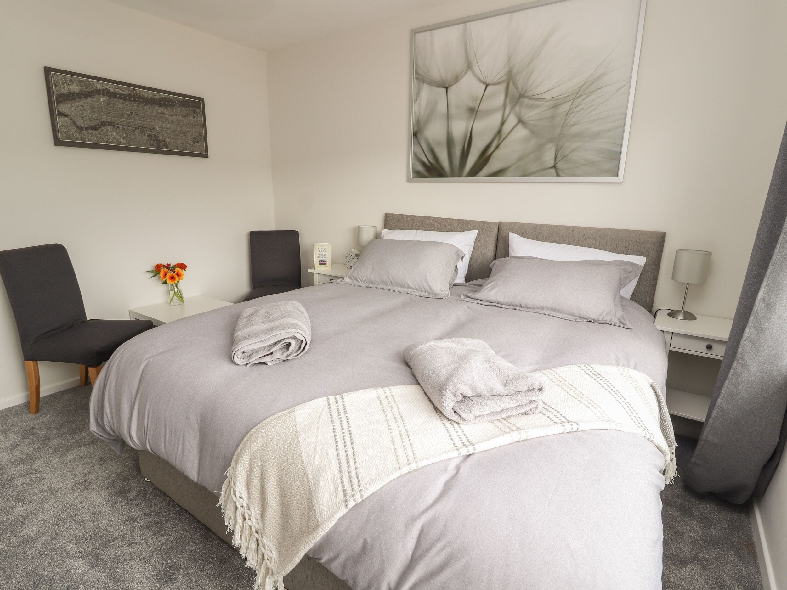 Una cama bien hecha con una manta gris y dos almohadas, una mesita con un florero, una silla y una obra de arte abstracto colgada en la pared.