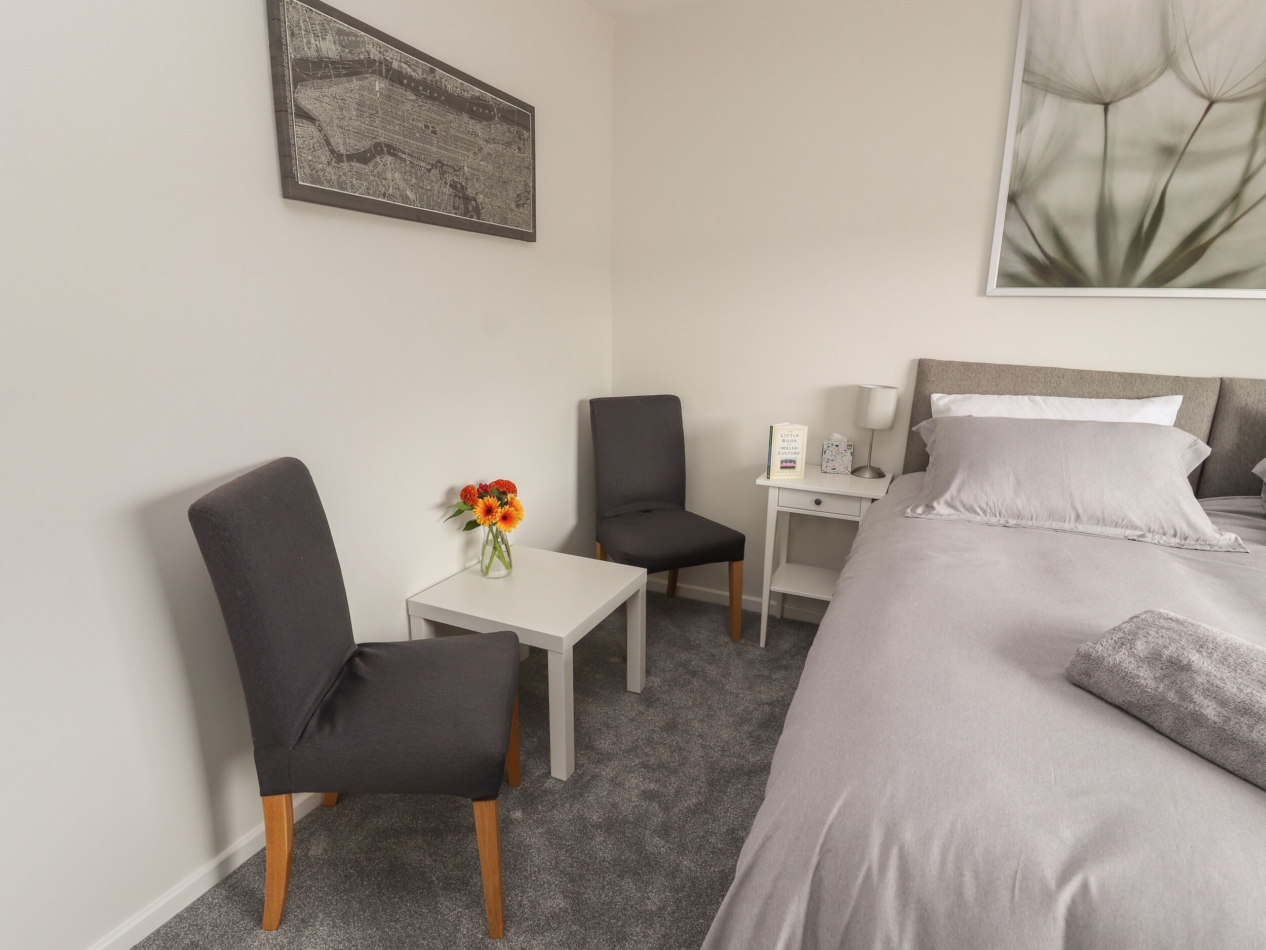 Un dormitorio con cama, dos sillones, una mesita con un jarrón de flores, una mesita de noche con lámpara y un cuadro en la pared.